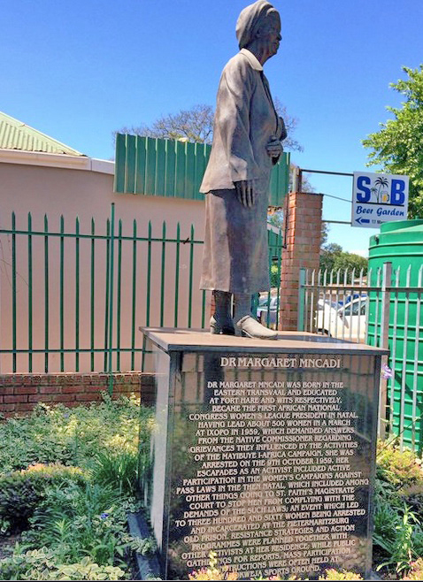 A brown granite statue of a woman wearing a calf-length skirt, jacket and beret, standing on a square plinth with an inscription on the side. The monument is in a garden with shrubs and lowers, bounded by a green palisade fence. A wall and a green water tank are visible through the fence.